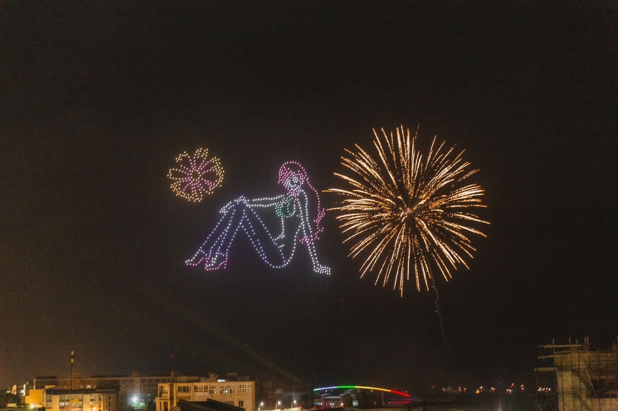 A nighttime view of a cityscape with a large illuminated figure of a person sitting on the ground, surrounded by fireworks.