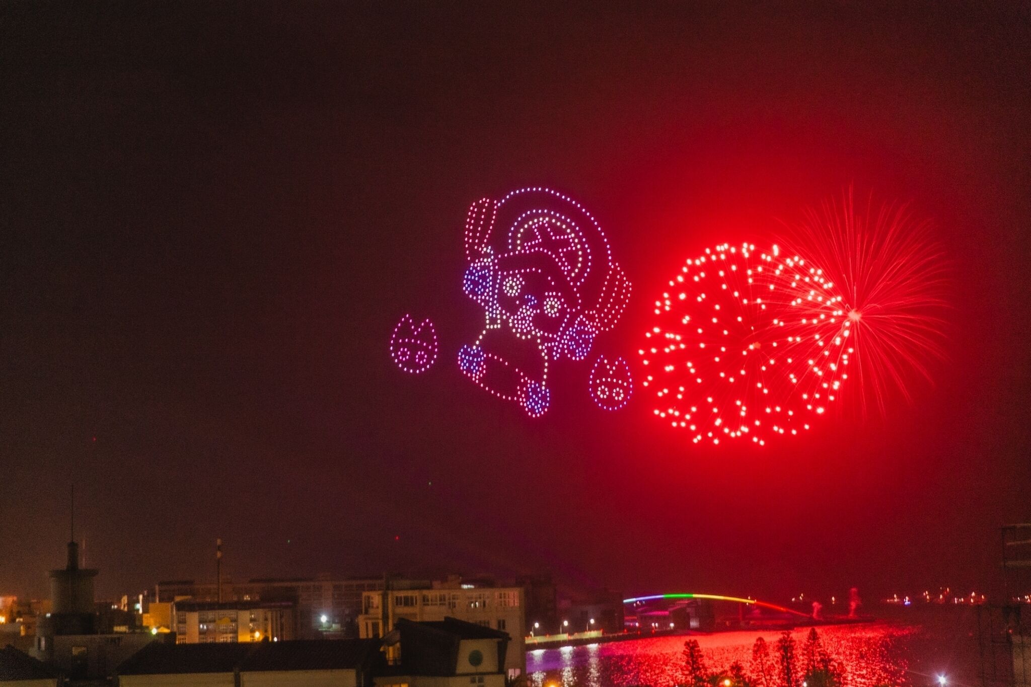 Yökuva kaupunkimaisemasta, jossa rakennukset on valaistu ja taivaalla näkyy suuri, värikäs valonäytös.