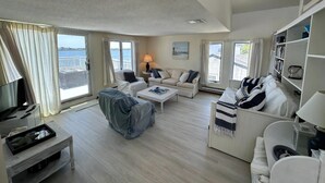 Ein geräumiges Wohnzimmer mit großem Fenster mit Blick auf ein Gewässer, inklusive Sofa, Couchtisch und Bücherregal.