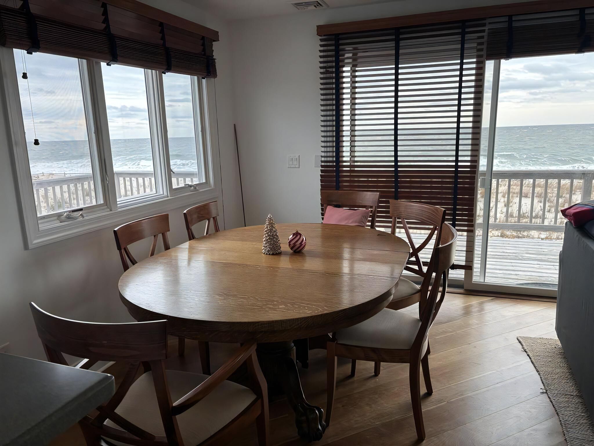 Una mesa de madera redonda con seis sillas, ventanas amplias con persianas y vista al océano.