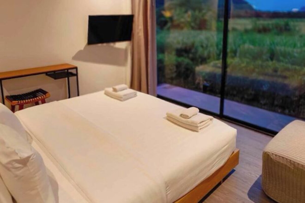 A hotel room with a large window offering a view of a field. There is a bed with white linens and a bedside table with a lamp.
