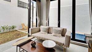 A modern living room with a sofa, a coffee table, and a view of a swimming pool through large windows. - Serila Villa by Kozystay - Jimbaran (Jimbaran)