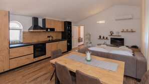A modern kitchen with wooden cabinets and a black range hood, a dining area with a wooden table and chairs, and a living area with a sofa and a TV.