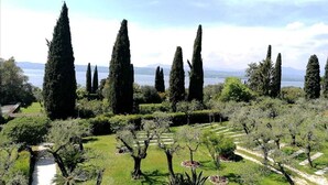 A landscaped garden with tall cypress trees, a variety of shrubs, and a clear view of a lake in the background. - Villa Paradiso (Sirmione)