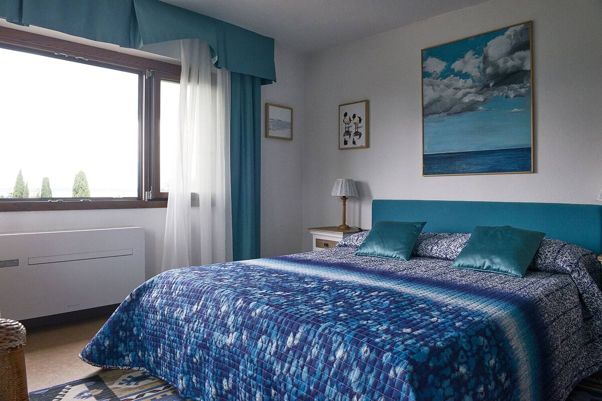 A bedroom with a large bed, a window with curtains, a nightstand with a lamp, and a painting on the wall.