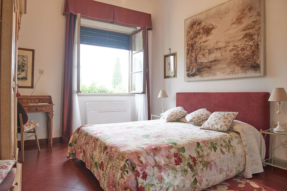 A bedroom with a bed, a desk, a chair, a window with curtains, and a painting on the wall.