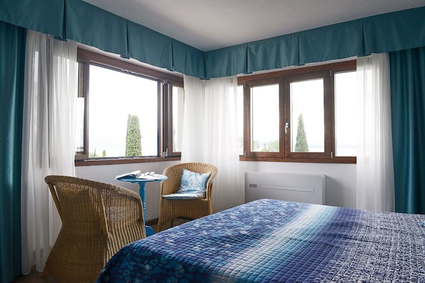 A bedroom with a bed, a chair, a small table, and two windows with curtains. - Villa Paradiso (Sirmione)