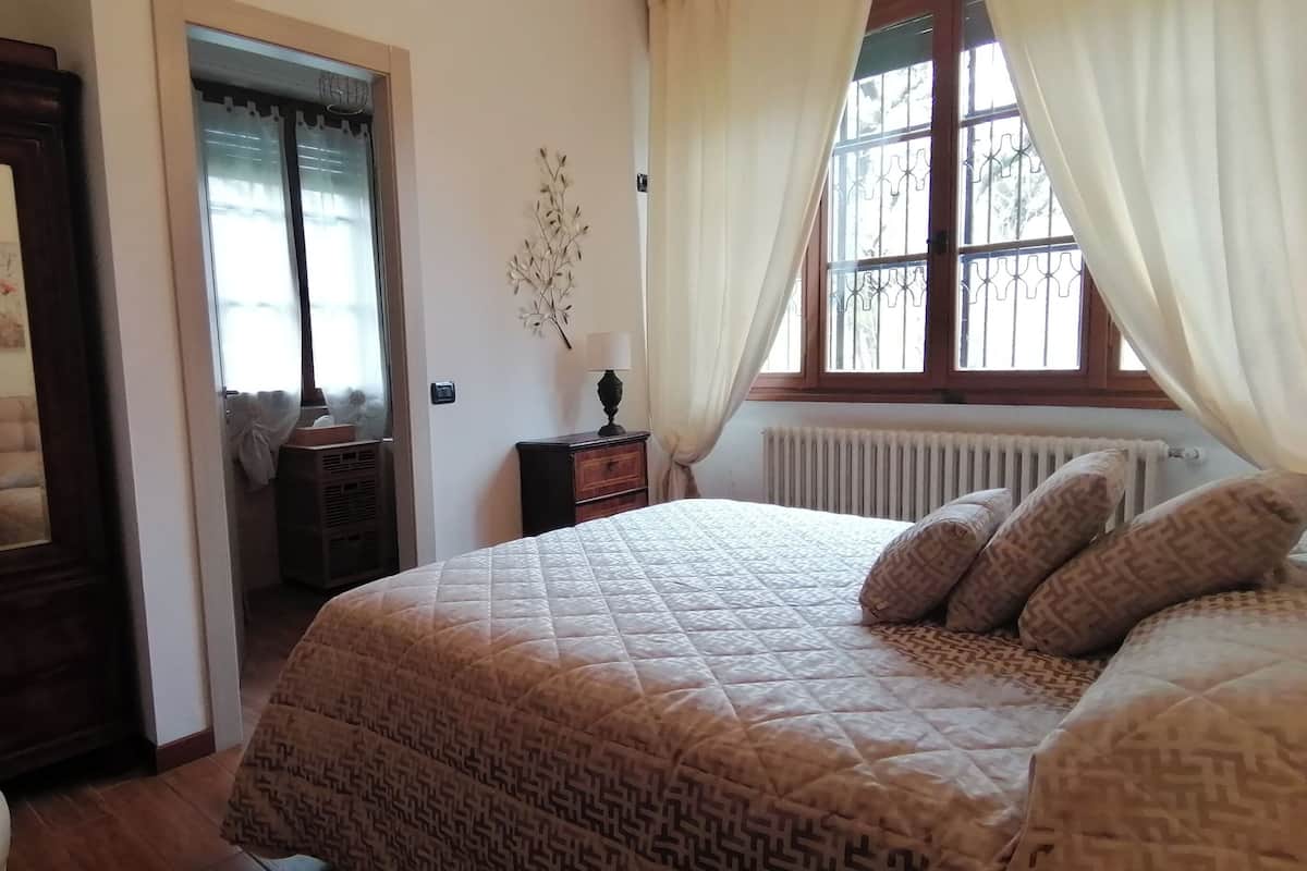 A bedroom with a large bed, a bedside table with a lamp, a radiator, and a window with curtains.