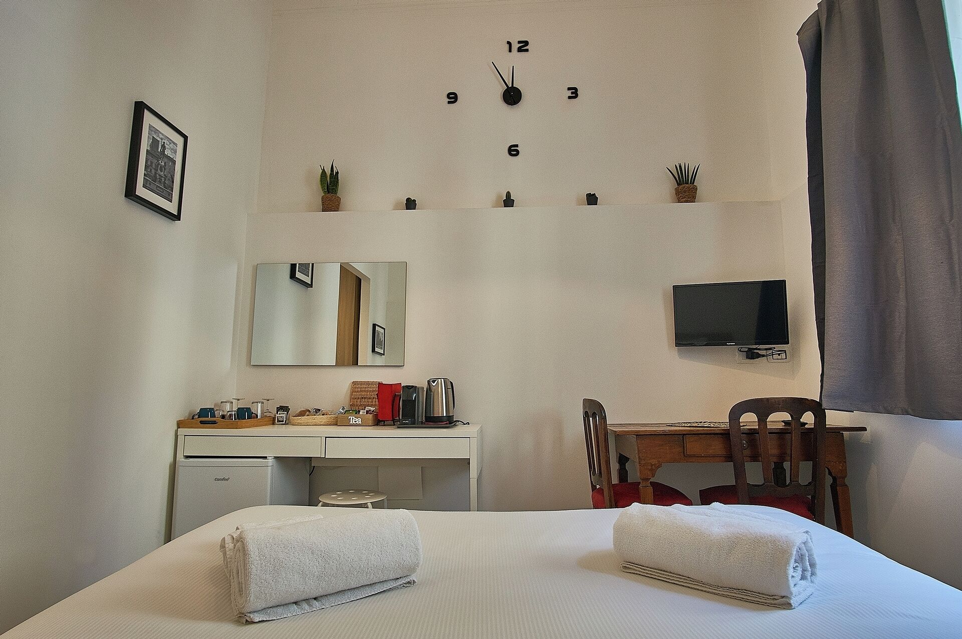 Una habitación de hotel con cama, escritorio, silla, televisor, espejo y un reloj en la pared.