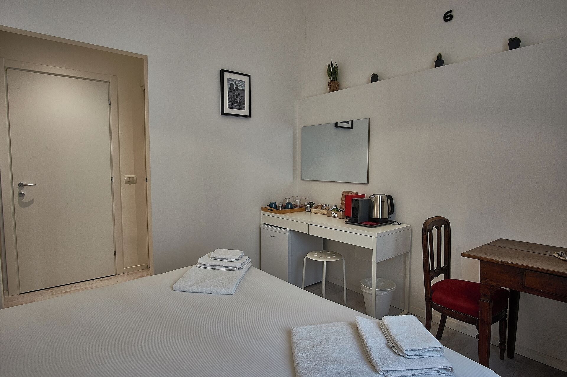 Ein Hotelzimmer mit einem Bett, einem Schreibtisch, einem Stuhl, einem kleinen Hocker, einem Spiegel, einer Ablage mit Dekoration und einer Tür.