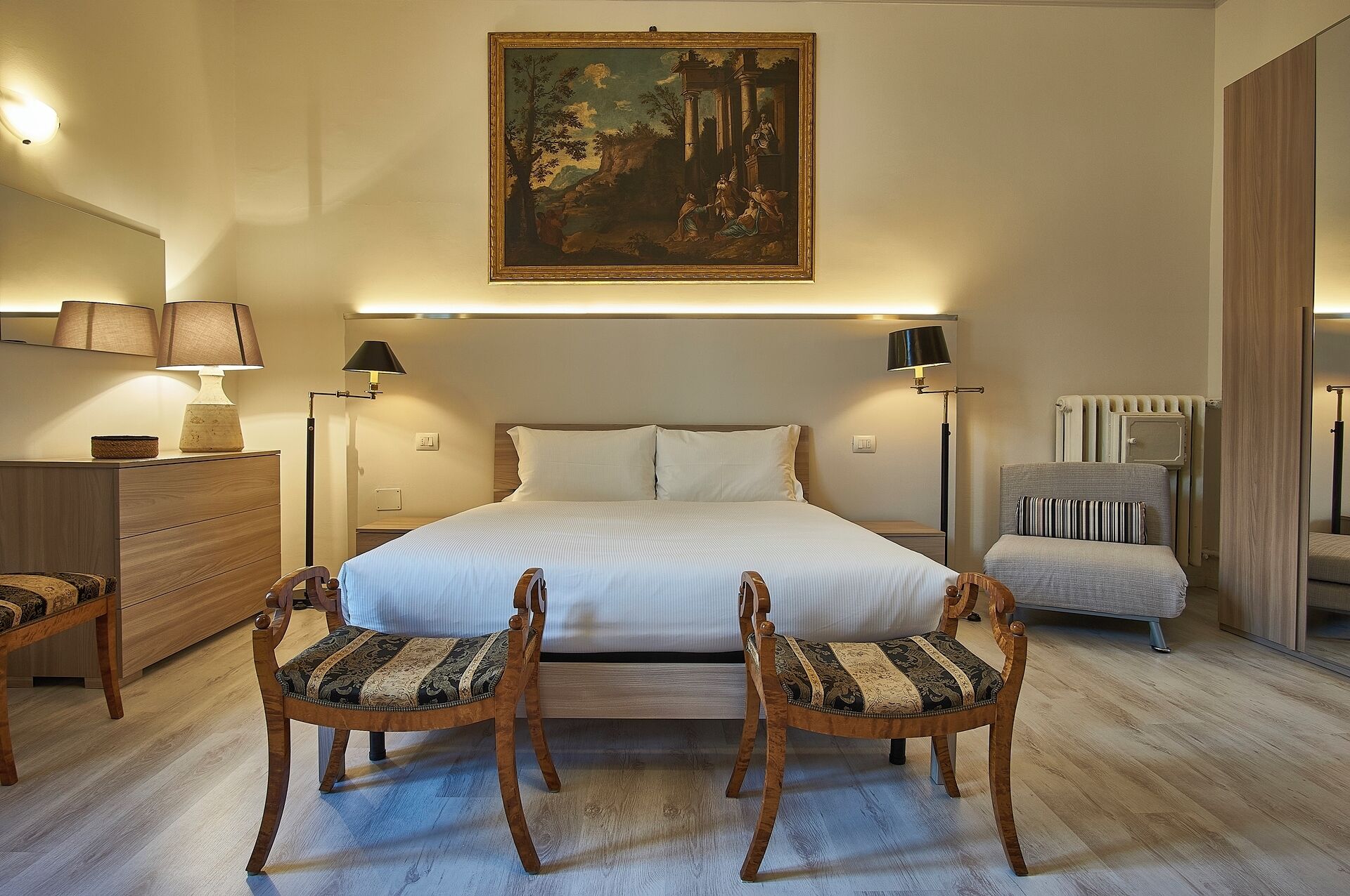 Una habitación de hotel con una cama grande, dos mesitas de noche, una butaca, una pequeña mesa y un cuadro en la pared.
