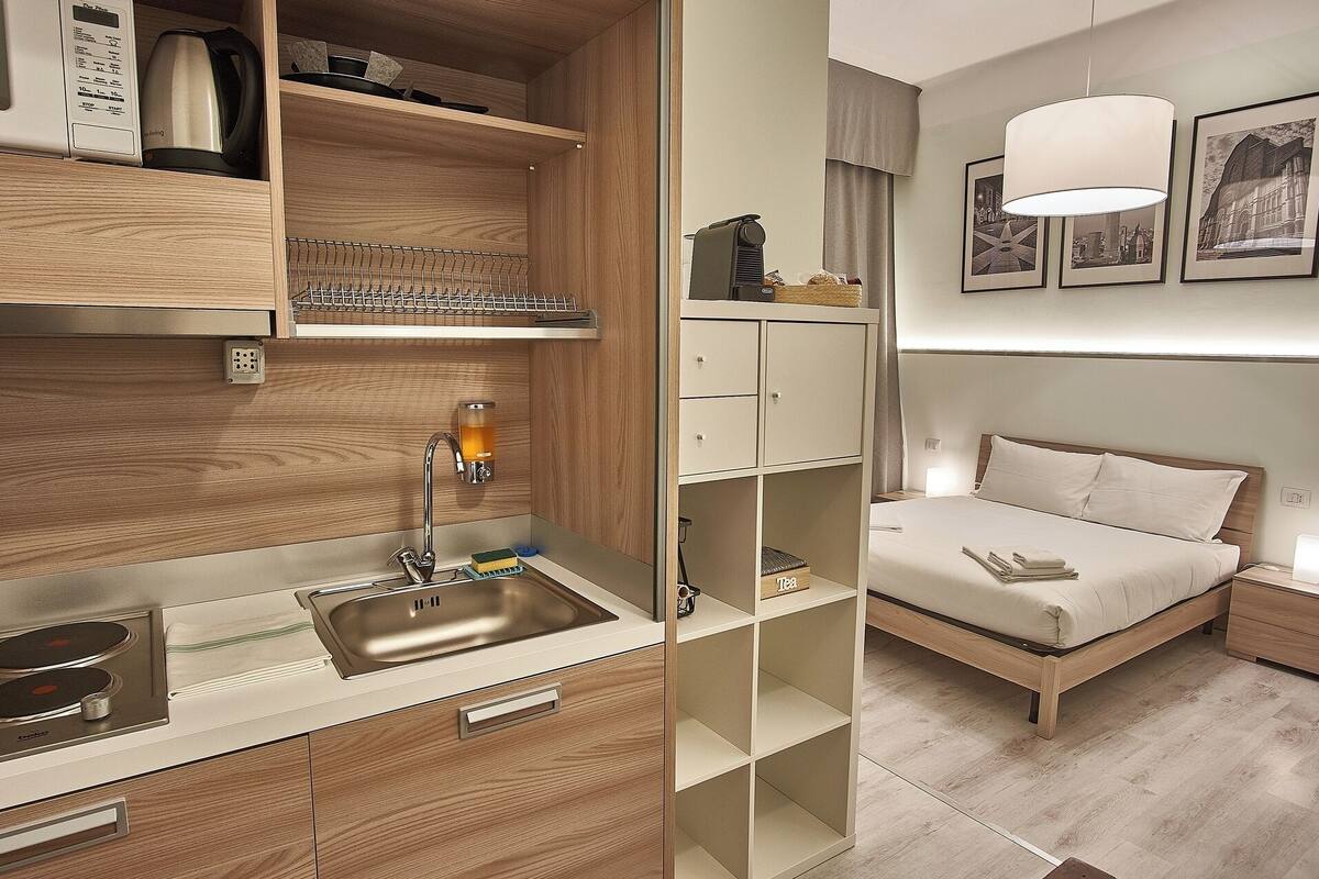 Ein modernes Hotelzimmer mit Küchenzeile, einem Bett mit Kopfteil, einem Nachttisch und einer Wandlampe.
