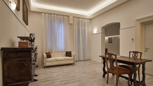 A room with a wooden dining table, two chairs, a sofa, a wooden cabinet, and a window with sheer curtains. - Cuore di Bologna Suites (Bologna)