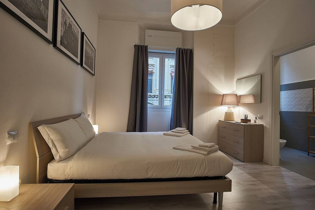 Ein Schlafzimmer mit Bett, Nachttischlampe, Kommode und Blick durch ein mit Vorhängen drapiertes Fenster hinaus.