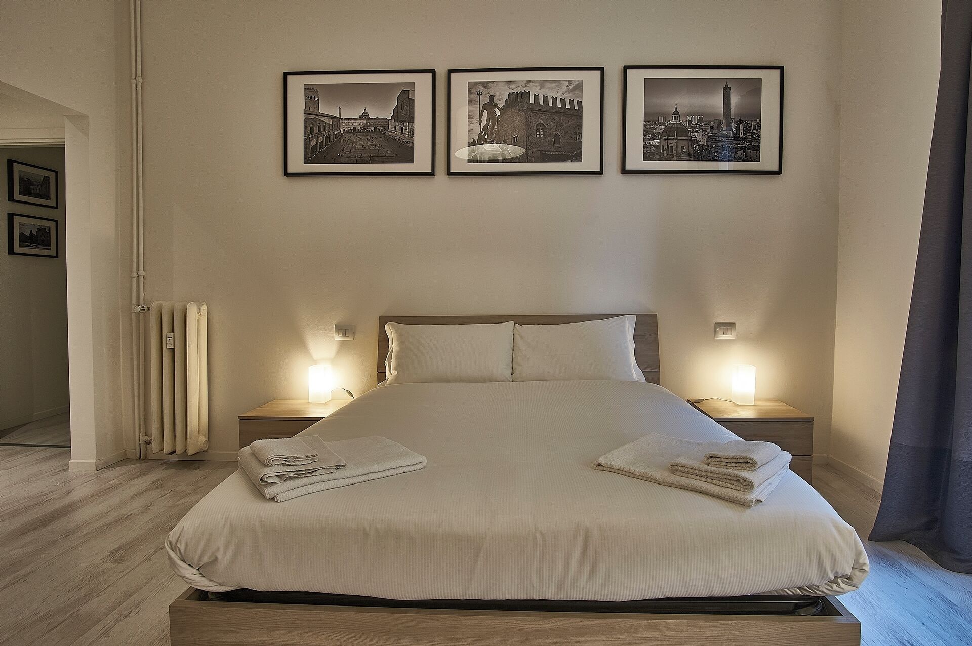 A neatly made bed with folded towels, a wooden headboard, and bedside lamps. Three framed pictures are hung on the wall above the bed.