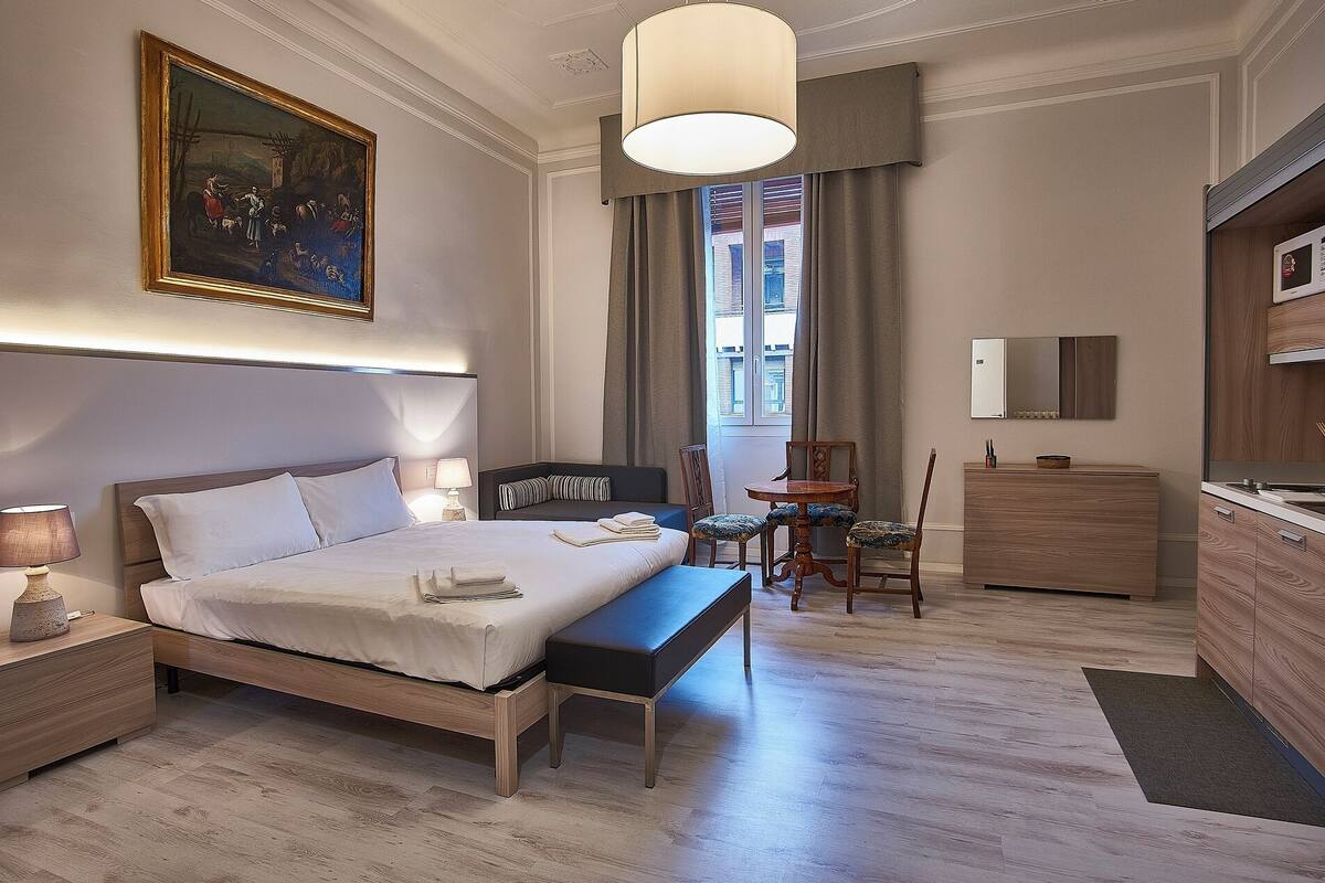 Ein Hotelzimmer mit einem großen Bett, einem Nachttisch, einer Lampe und einem Bild an der Wand. Es gibt einen kleinen Tisch mit Stühlen sowie eine Holzkommode mit Spiegel.