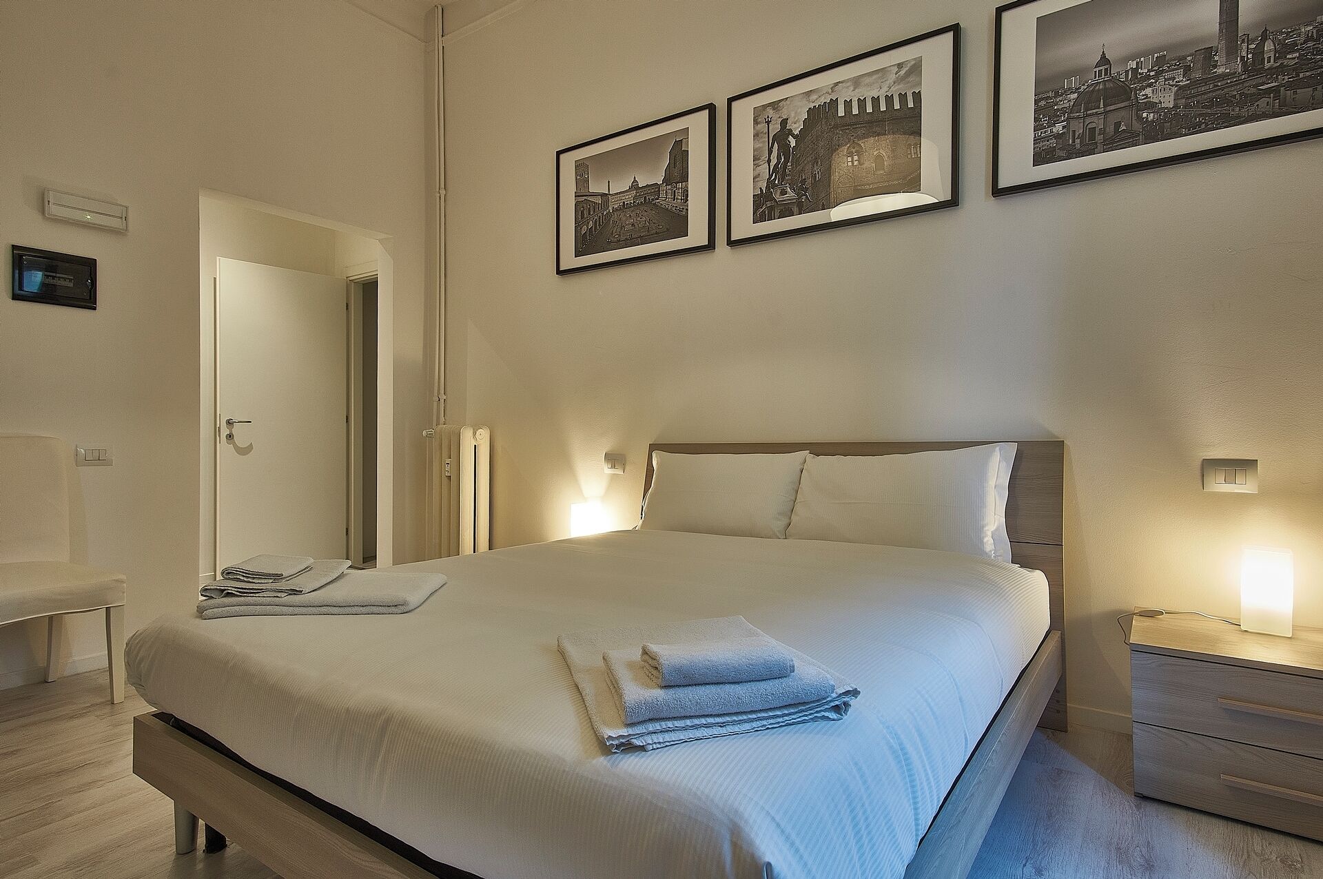 Un lit bien fait, avec des serviettes pliées, une table de chevet avec une lampe et trois tableaux encadrés accrochés au mur.