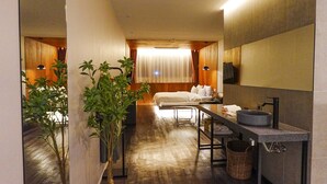 Habitación de hotel con cama, escritorio, lavamanos y un espejo grande. Hay plantas y una ventana con cortinas.