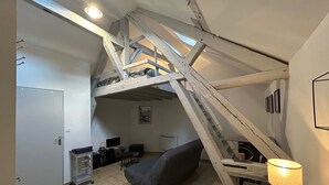 Une chambre à coucher aménagée dans un espace sous les toits, avec un plafond en pente, un canapé gris, un bureau avec une lampe et un escalier.