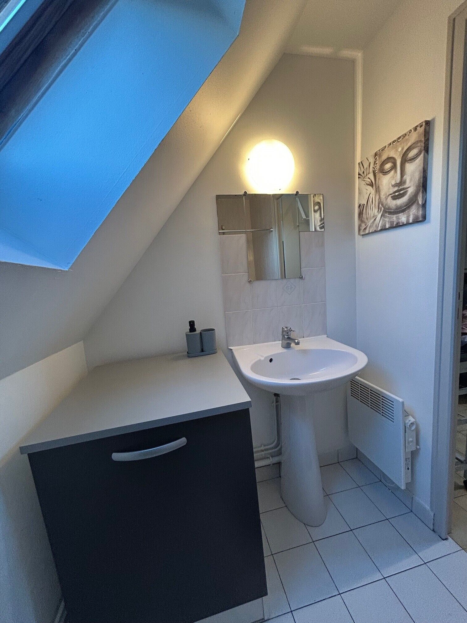 Une salle de bain avec un lavabo blanc, un miroir et un porte-serviettes. Il y a une fenêtre avec un cadre bleu et un luminaire au-dessus du miroir.