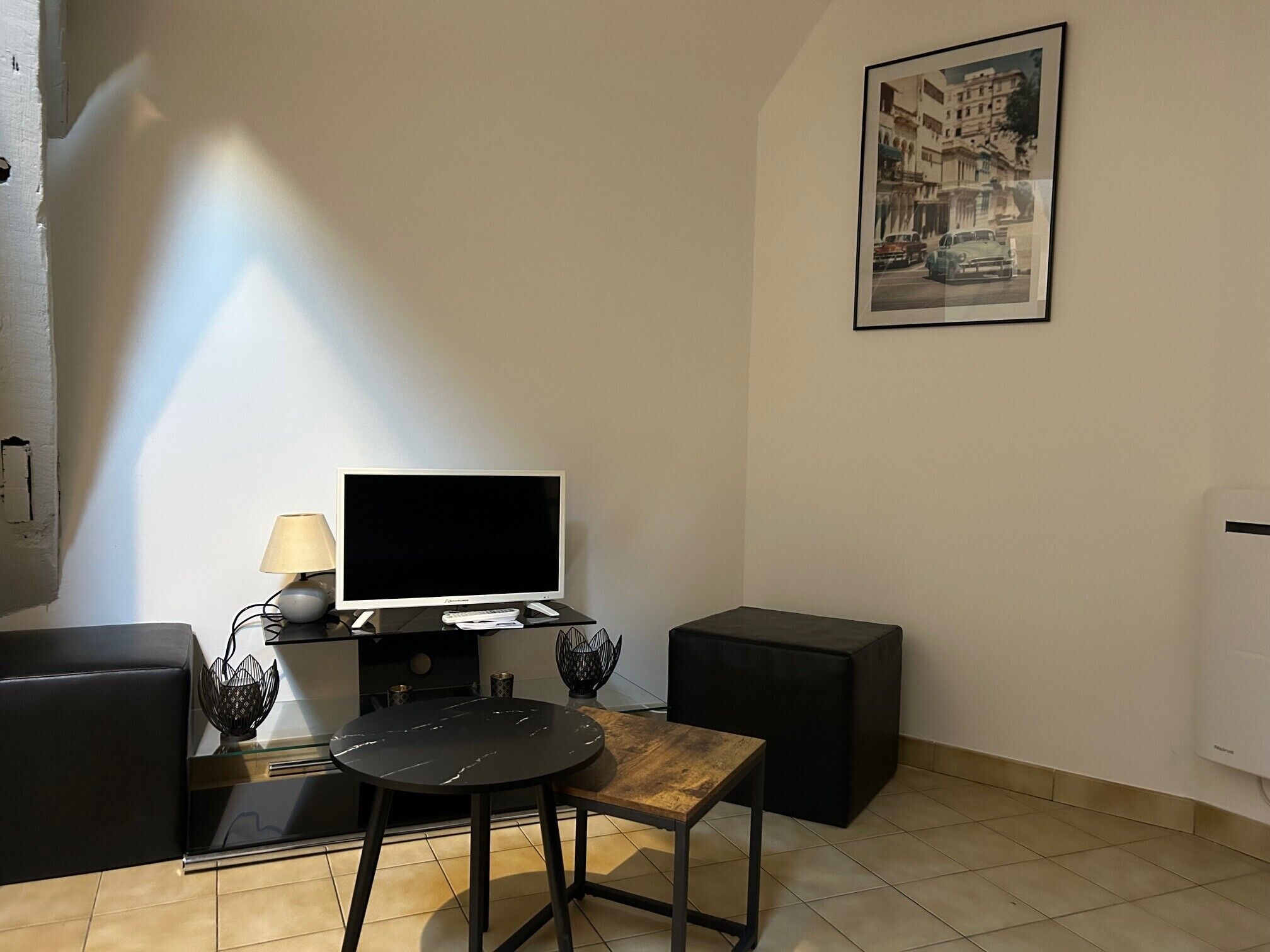 Un petit salon moderne doté d’un téléviseur à écran plat, d’une table basse en bois, d’un canapé noir et d’un pouf noir. Un tableau encadré orne le mur, et une lampe est posée sur la table d’appoint.