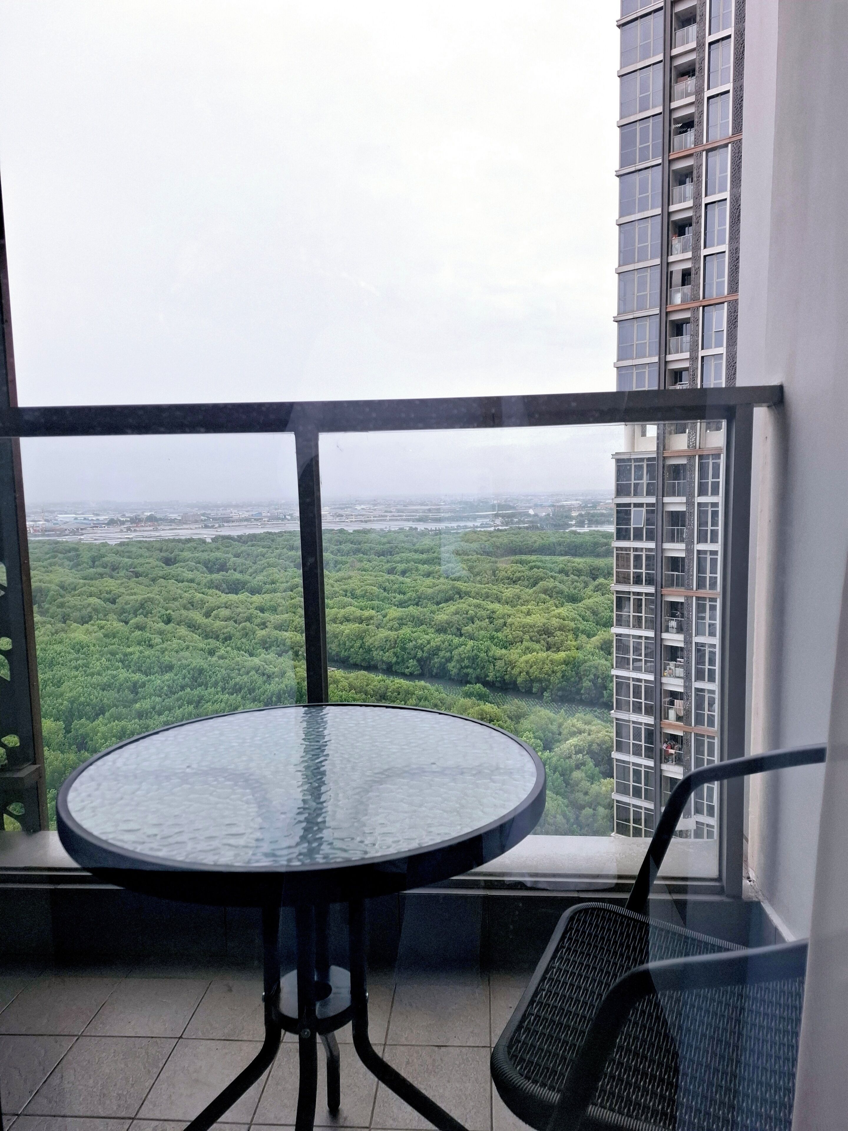 A balcony with a round glass table and metal chairs, overlooking a dense green forest and a cityscape.