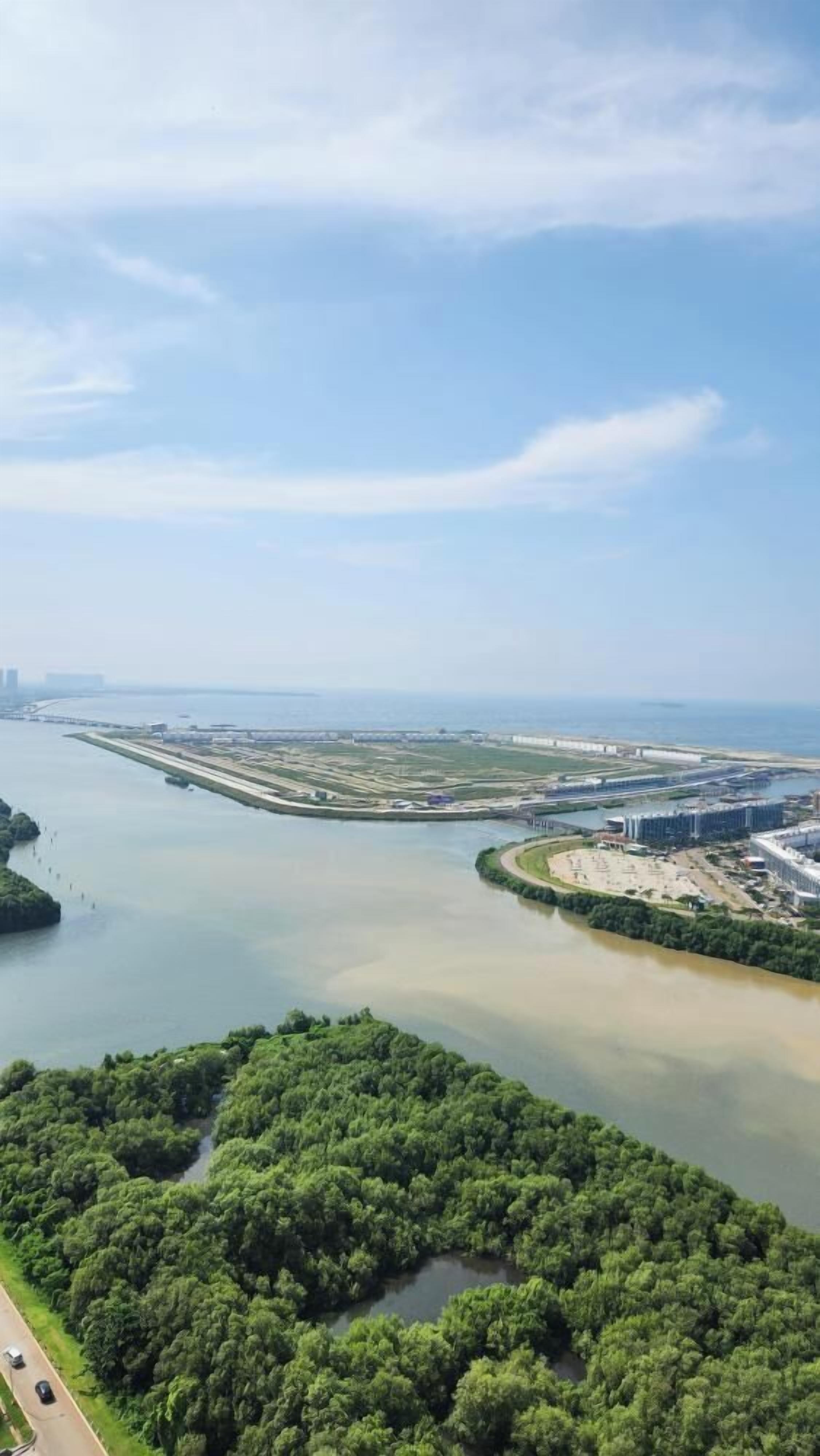 An aerial view of a coastal area with a river, green forested areas, and a developed waterfront with buildings and infrastructure.
