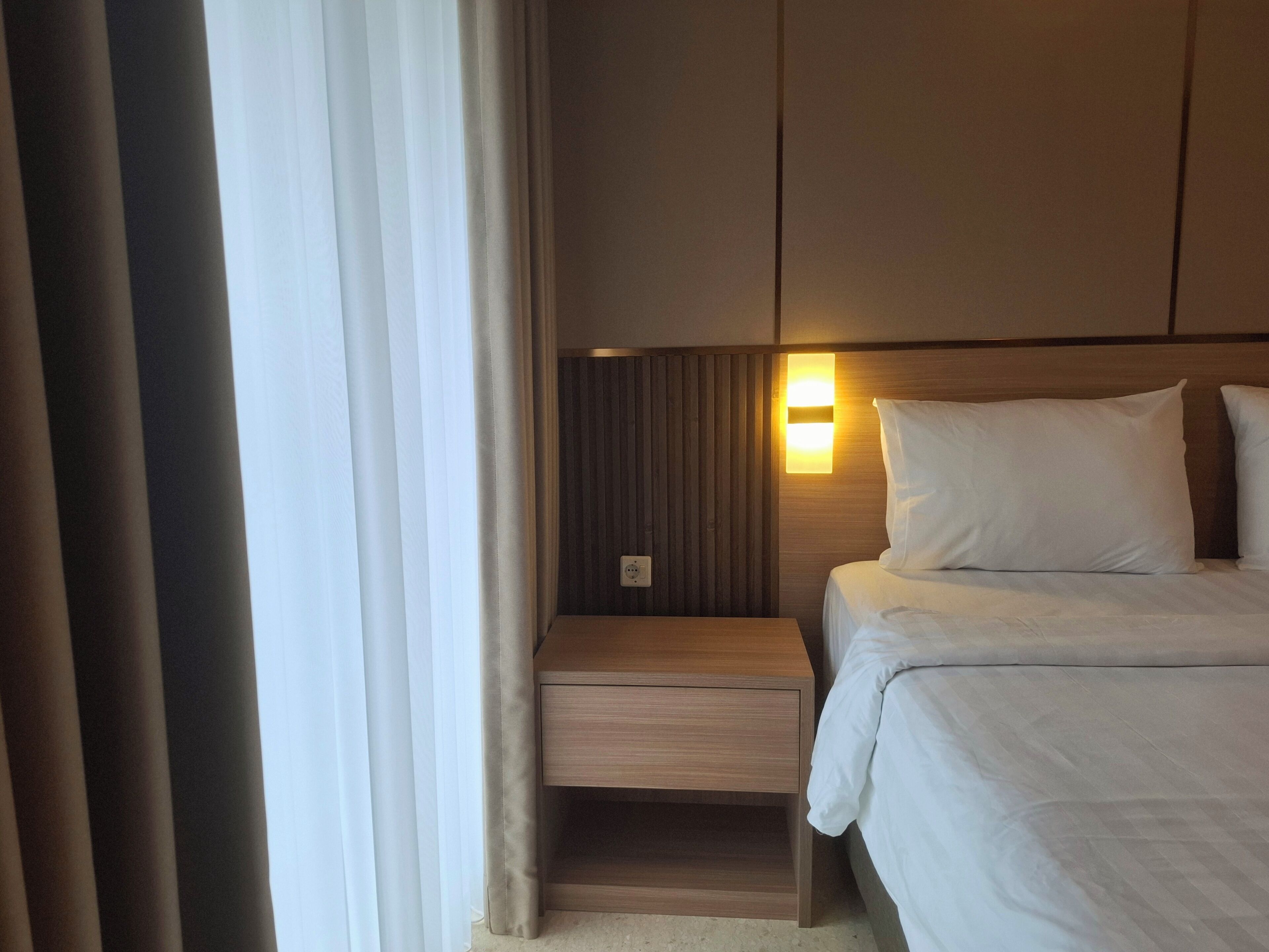 Hotellihuone, jossa on sänky, yöpöytä, seinään kiinnitetty valaisin ja ikkuna, jossa on verhot.