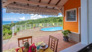 Un espacio exterior cubierto con piscina, sillas y una mesa lista para comer, con vistas a un paisaje pintoresco con colinas y el océano.
