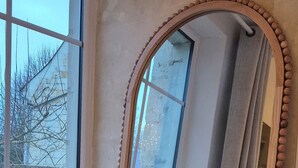 Ein großer Spiegel mit Holzrahmen, neben einem Fenster mit Blick auf kahle Bäume, und zwei weiße Vasen mit getrockneten Blumen auf dem Boden.