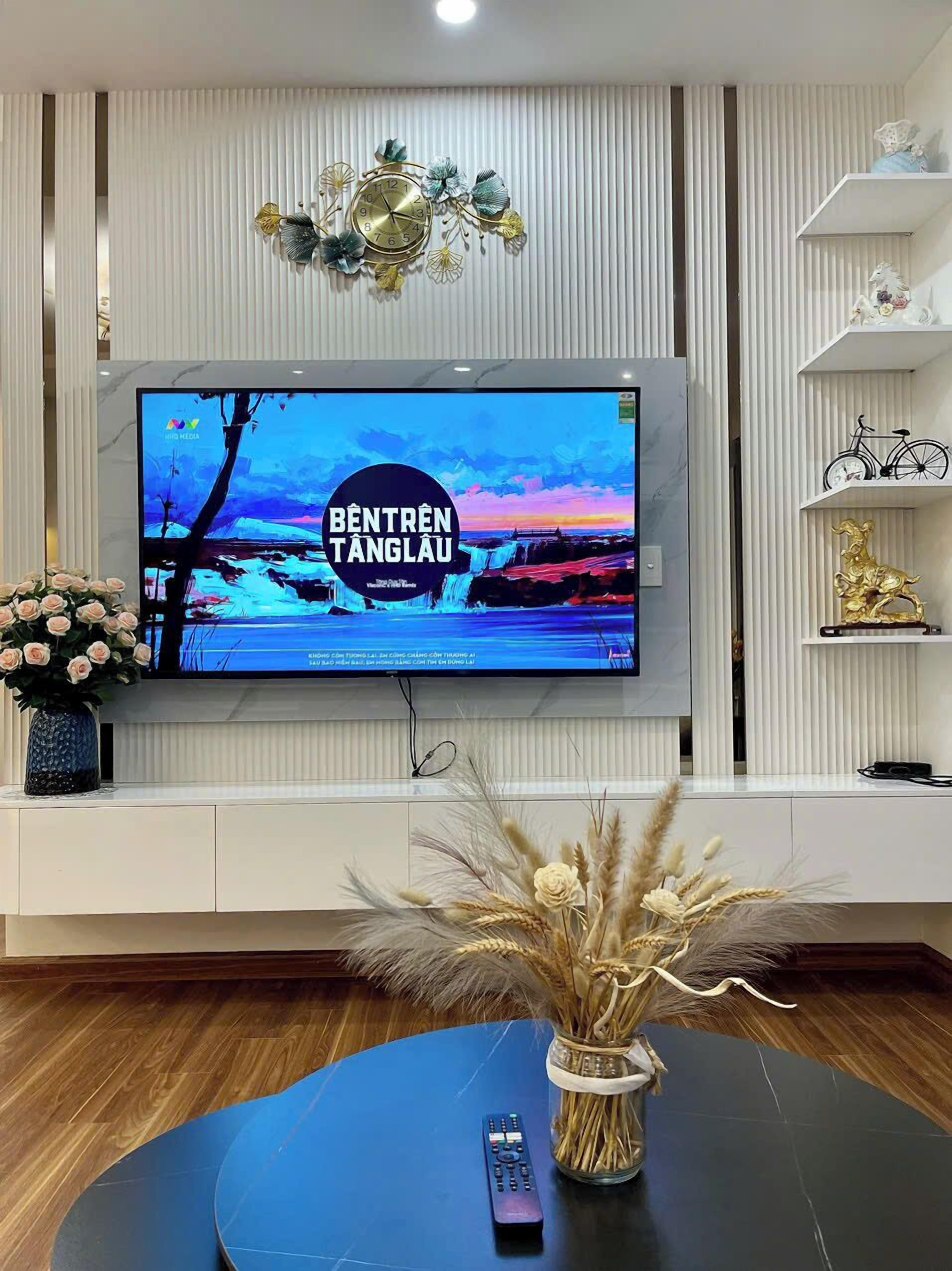 Ein modernes Wohnzimmer mit einem flachbildfernseher an einer weißen Wand, einer dekorativen Uhr und einer Vase mit Trockenblumen auf einem blauen Tisch.