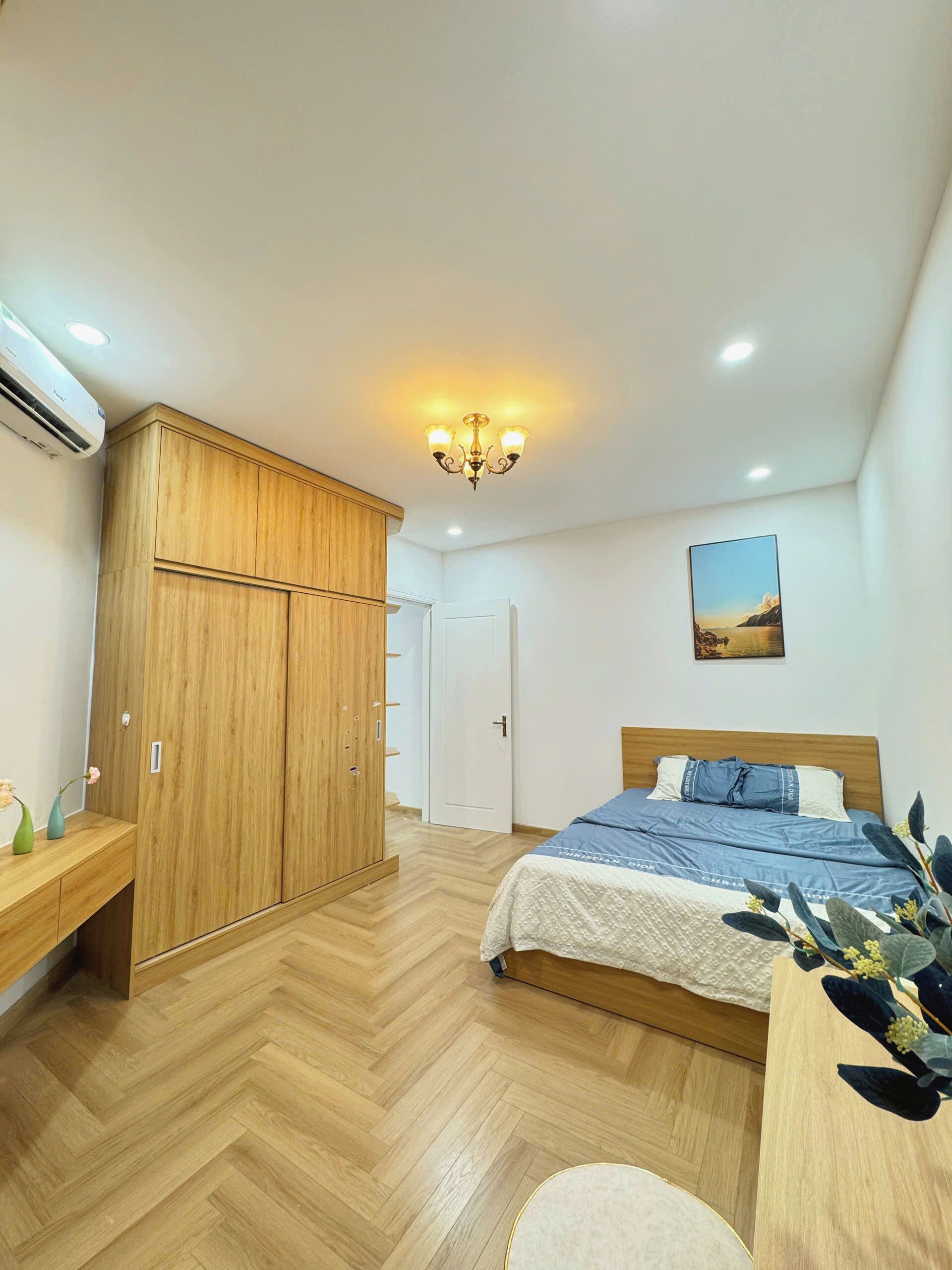 A bedroom with a wooden wardrobe, a bed with a blue bedspread, a white bedside table, and a wall-mounted picture.