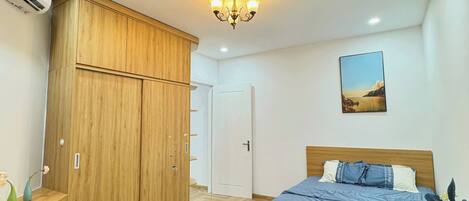 A bedroom with a wooden wardrobe, a bed with a blue bedspread, a white bedside table, and a wall-mounted picture.