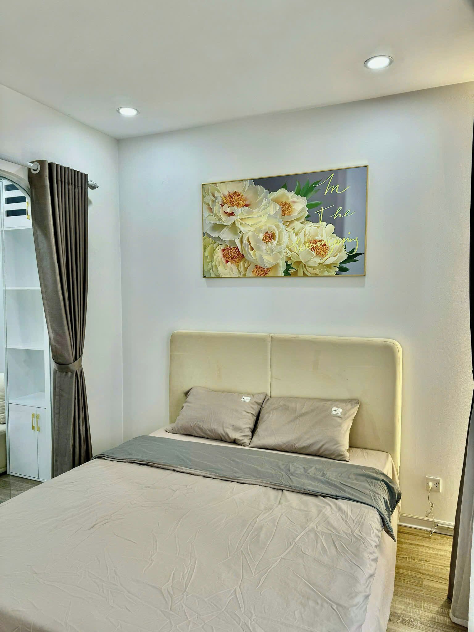 A bedroom with a bed, a headboard, a painting of flowers, a wardrobe, and a curtain.