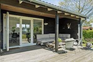 A wooden deck with a sofa and chairs, a dining table, and a sliding glass door leading to an indoor area with a desk and chairs.