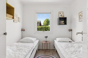A bedroom with two beds, a small table, a window with a view of a house and trees, and a shelf with books and a plant.