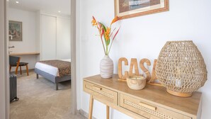 Una habitación de hotel con cama, escritorio, silla, un florero con flores, una consola de madera con adornos y un cuadro en la pared.