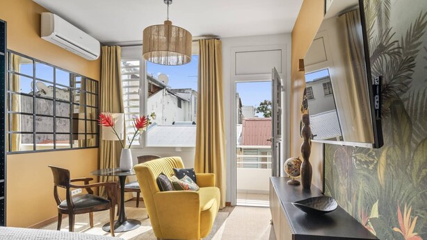 Una camera d'albergo con un letto, una poltrona gialla, un tavolino con un vaso, una televisione a parete e un balcone con vista sugli edifici.
