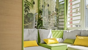 Ein grünes Sofa mit gelben und weißen Kissen, ein kleiner Tisch und ein Fenster mit Blick auf Gebäude und Pflanzen.