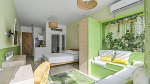 Ein modernes, helles Zimmer mit Bett, Sofa und Essbereich. Es gibt ein großes Fenster mit Pflanzen, eine Pendelleuchte und eine Klimaanlage.