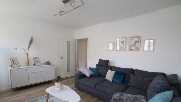 Een woonkamer met een grijze bank, een witte salontafel, een kamerkleed met dessin, een witte kast met ingelijste foto's, en een deur die leidt naar een andere kamer.