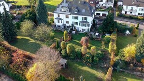 Een groot, tweelaags huis met een schuin dak, omringd door een verzorgd gazon en verschillende bomen.