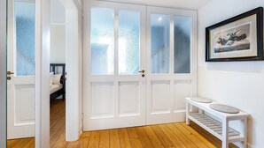 Een kamer met een houten vloer, een witte bank met kussens, een witte deur met geëtste glaspanelen, en een ingelijst schilderij aan de muur.