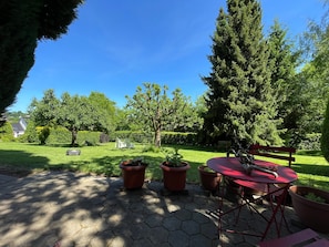 Eine Terrasse mit einem roten Tisch und Stühlen, umgeben von Topfpflanzen und einem großen Baum.