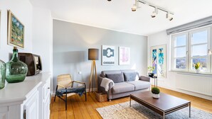 Een moderne woonkamer met een grijze bank, een houten salontafel, een staande lamp en uitzicht door grote ramen.