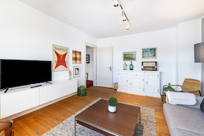 Ein Wohnzimmer mit einem Flachbildfernseher, weißen Schränken, einem braunen Couchtisch, einem grauen Sofa und einem Korbstuhl.