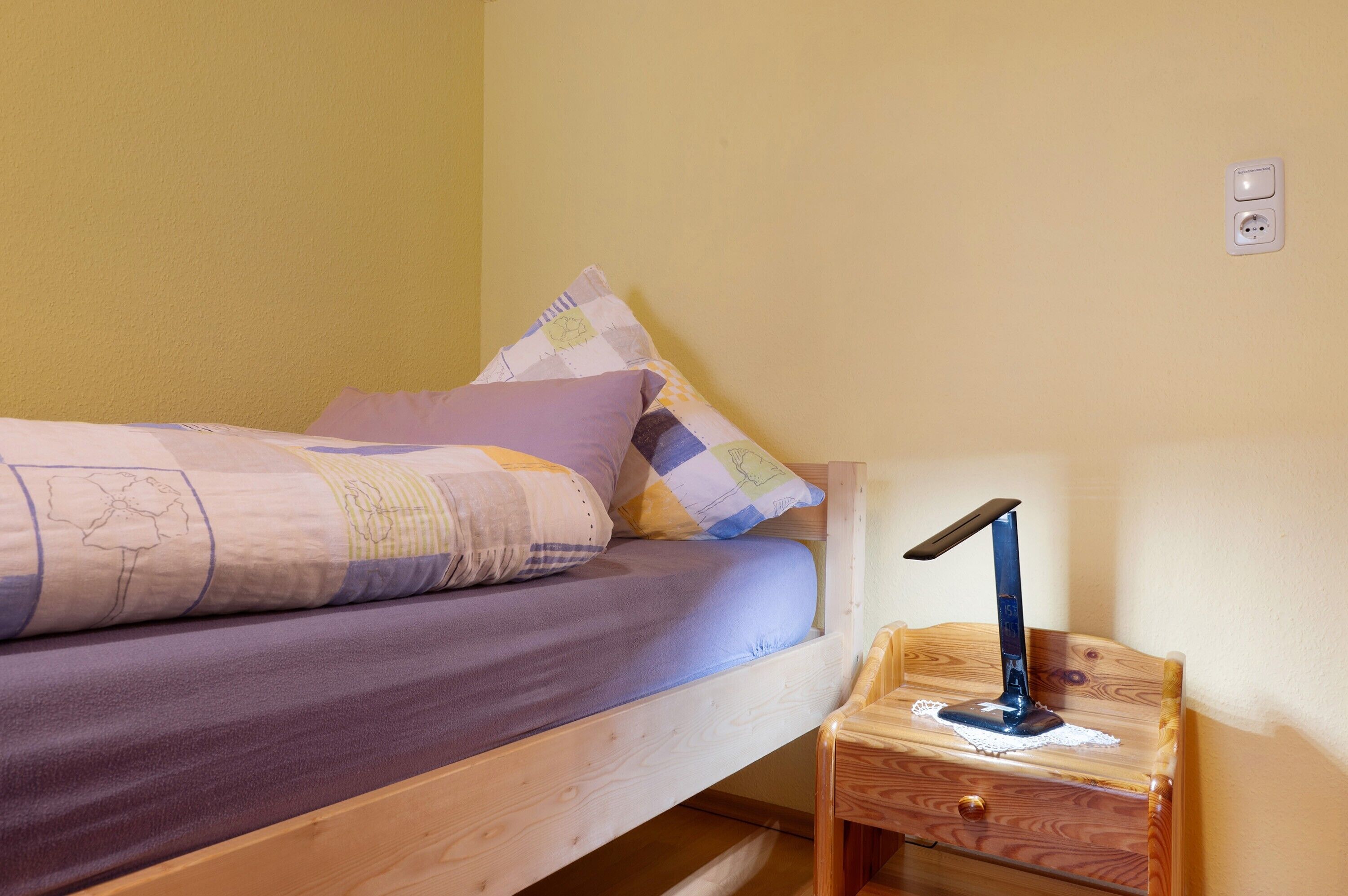 Una cama individual con estructura de madera, una mesita de noche con lámpara y un enchufe de pared.