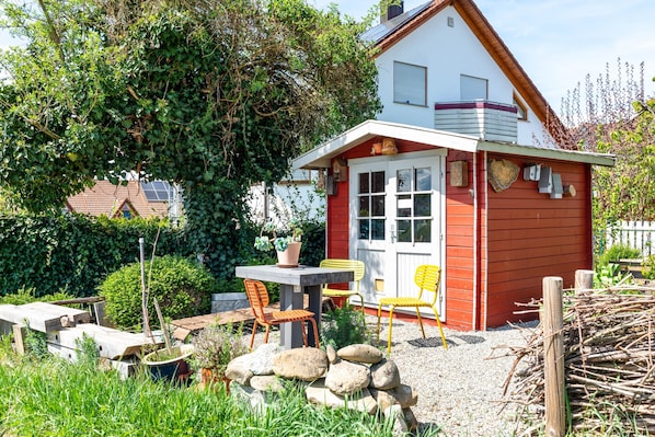 Une petite maison en bois rouge avec une porte blanche, une table et deux chaises, le tout situé dans un jardin.