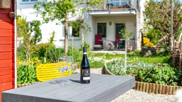 A table with a bottle of wine and two glasses, set outdoors with a garden and a building in the background.