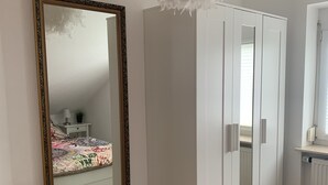 Une chambre avec un grand miroir, une armoire, un lit recouvert d’une couvre-lit à motifs floraux et une petite table de chevet avec une lampe.